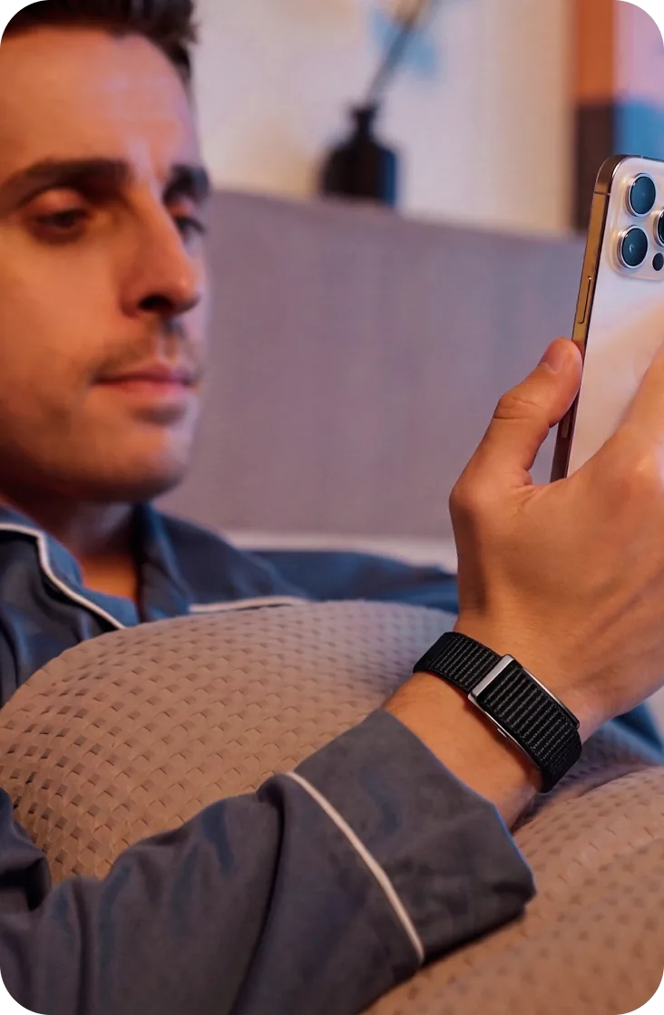 A man in bed checking his phone, wearing the HLTH Band