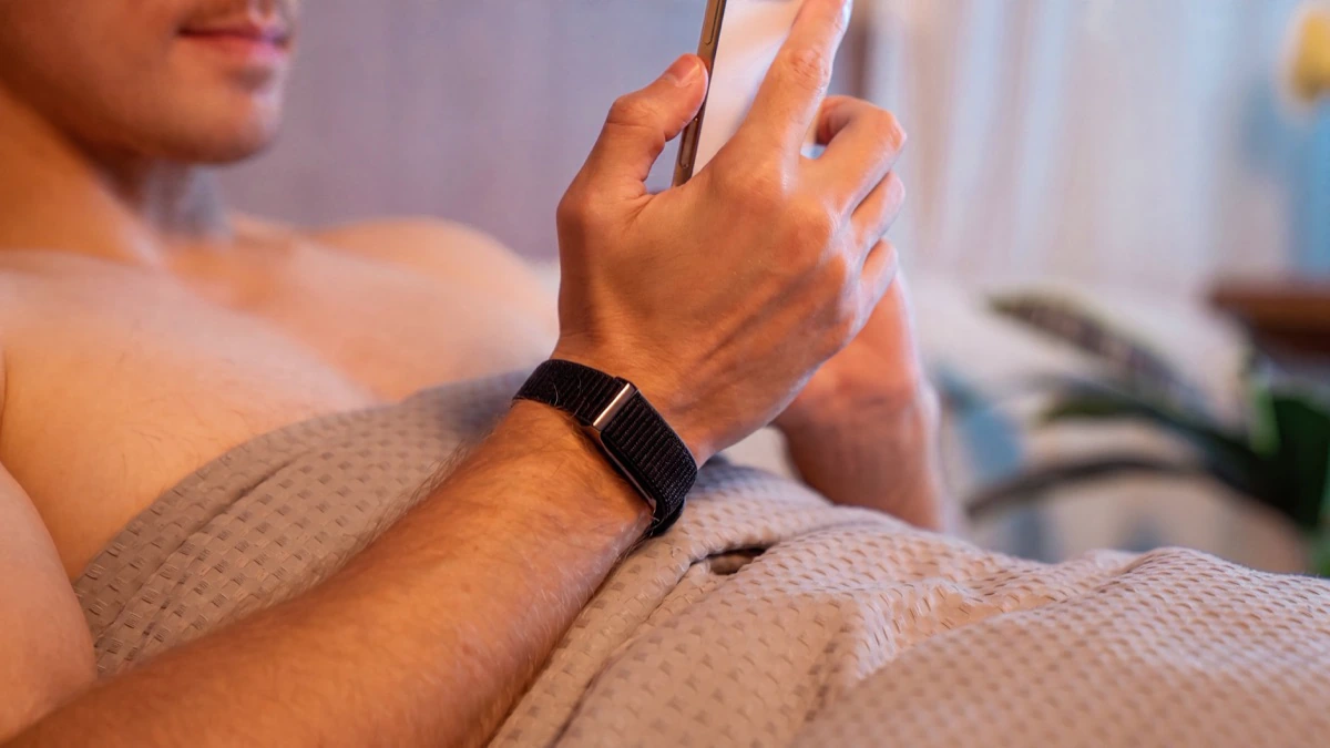 A man propped up in bed looking at his phone, the HLTH Band on his wrist