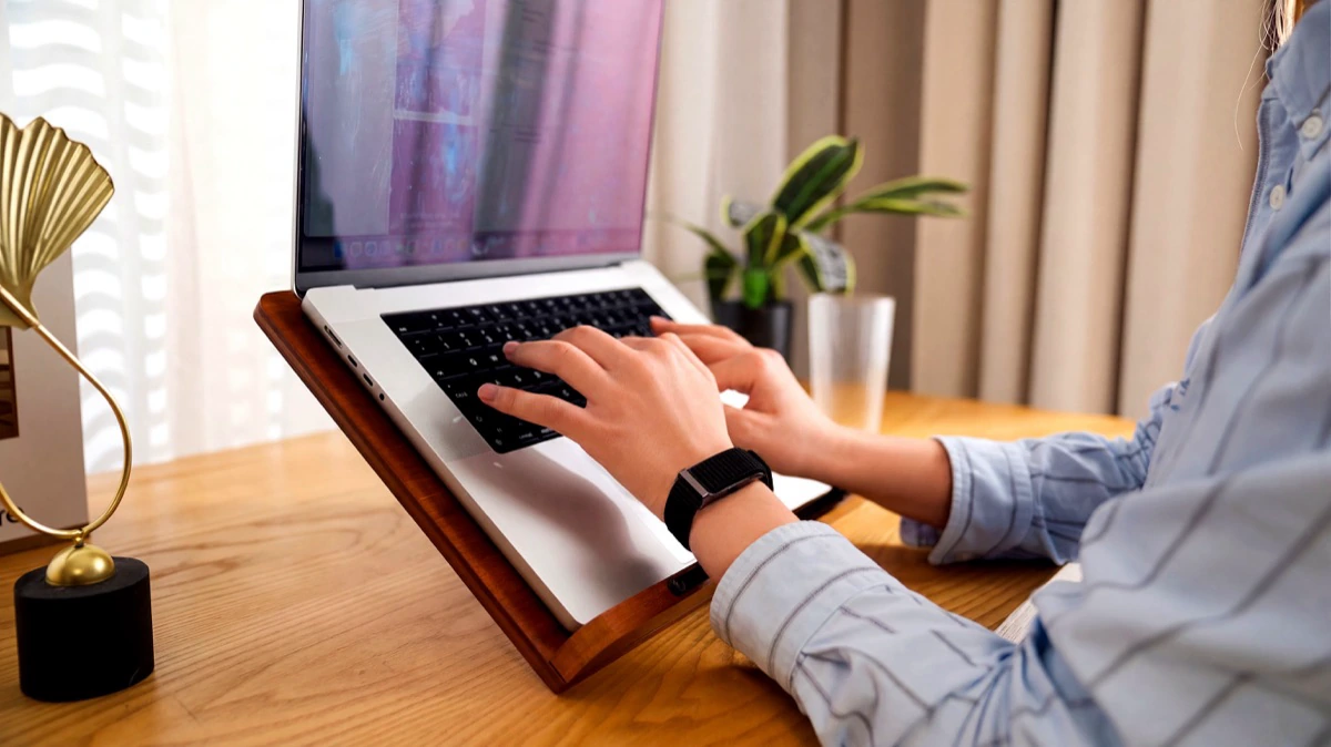 A woman typing at a laptop on a wooden desk, the HLTH Band on her wrist