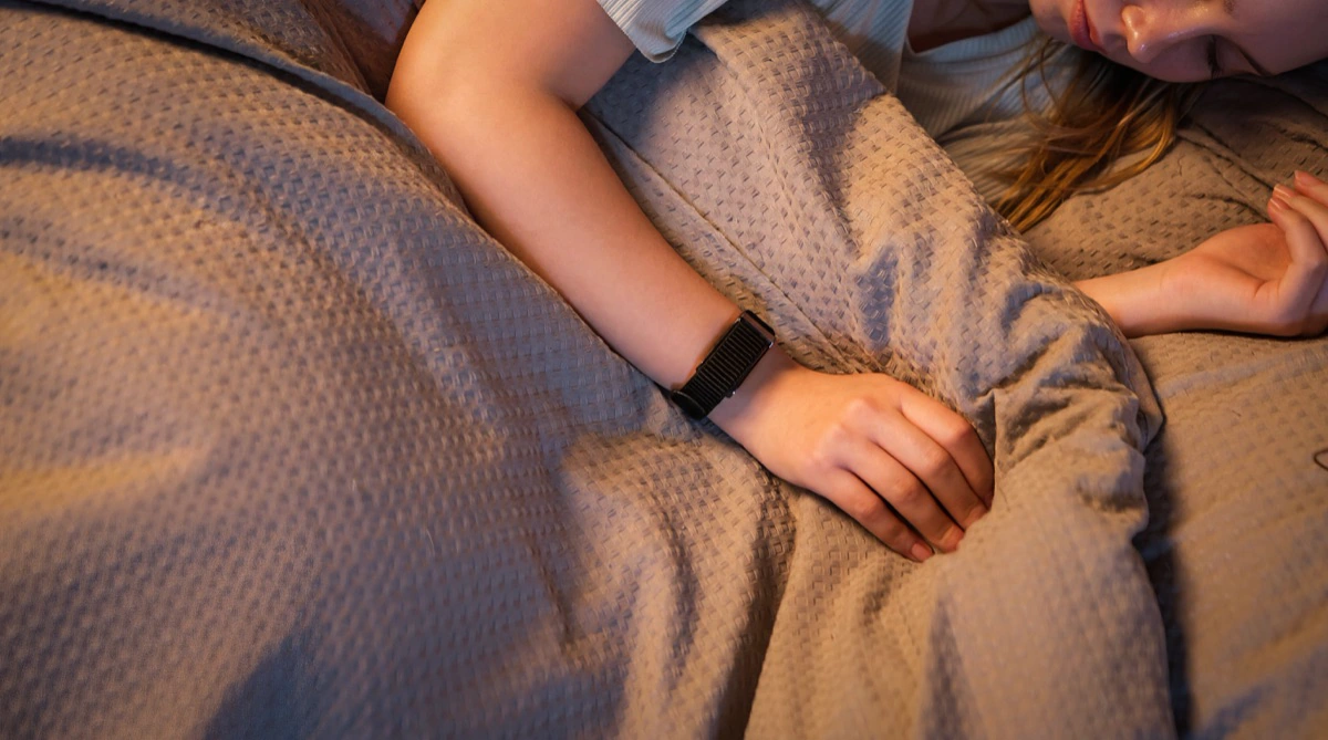 A sleeper's arm resting on a duvet in low lamplight, the HLTH Band on the wrist